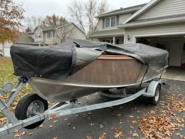 Aluminium sloep op boottrailer in oprit, gedeeltelijk bedekt met winterzeil, herfstbladeren rondom wielen