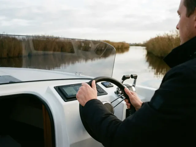Handen aan het stuur van elektrische boot op kalme Nederlandse waterweg met rietoevers in zacht ochtendlicht