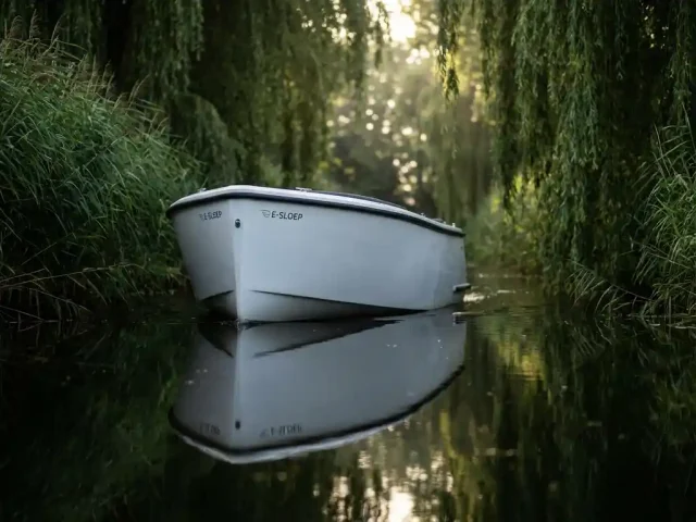 Witte elektrische sloep vaart door smalle gracht omringd door riet en hangende wilgentakken in ochtendlicht