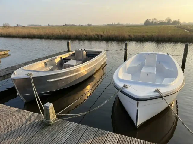 Twee sloepen, aluminium en polyester, afgemeerd aan houten steiger op Nederlandse waterweg met rietoevers