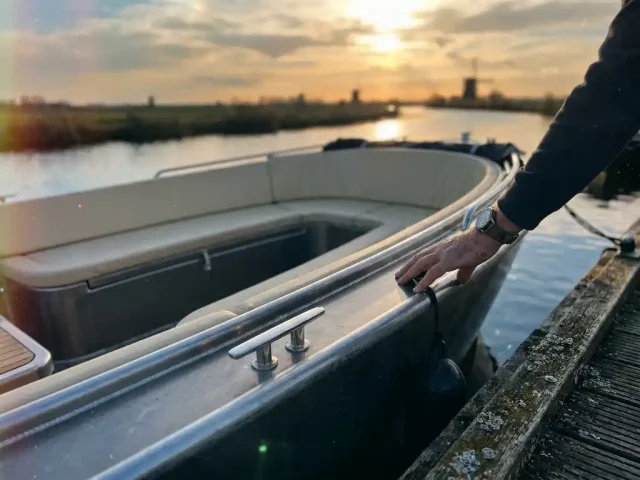 Aluminium tender boot afgemeerd aan houten steiger, hand op de dolboord, polderwater met gouden weerspiegeling en crèmekleurige kussens zichtbaar.