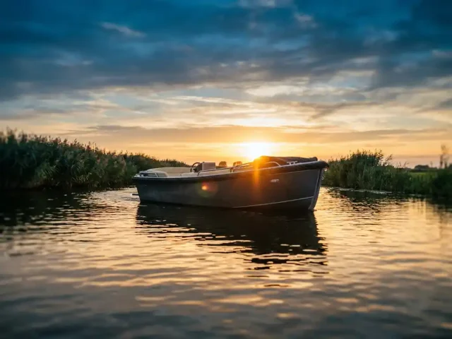 Antracietgrijze aluminium tender op rustig polderwater tijdens gouden uur, met warme reflecties en rietkragen op de achtergrond.