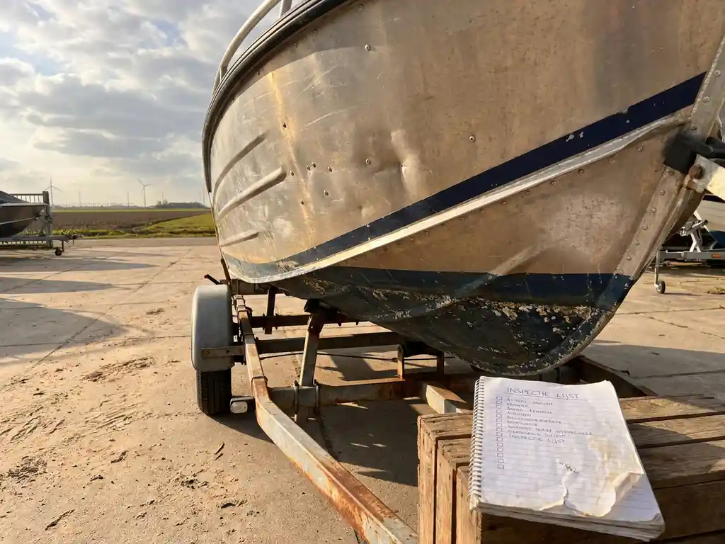 Verweerde aluminium tenderscheepsromp op trailer in dealeryard, met zichtbare klinknagelpanelen en verouderde waterlijnsporen in middaglicht.