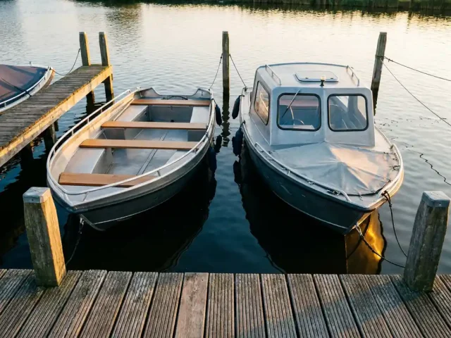 Twee aluminium sloepen afgemeerd aan een verweerde houten steiger in de polder, met kalm water dat goudkleurig middaglicht weerkaatst.