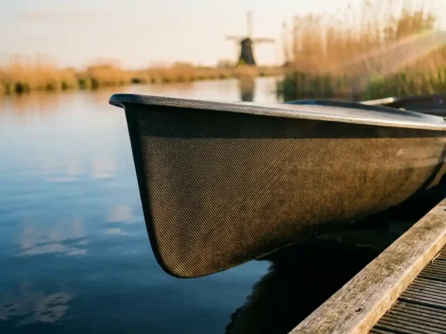 Koolstofvezel tender rompvlak met zichtbaar weefselpatroon op houten steiger langs rustig polderwaterland bij gouden uur.