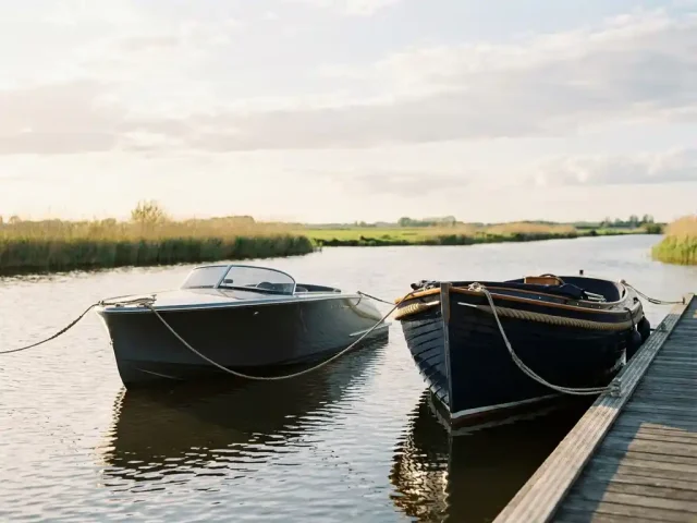Twee boten afgemeerd aan een houten steiger in de polder, een sportieve lage tender naast een klassieke hoge tender, in kalm water met rietkraag.