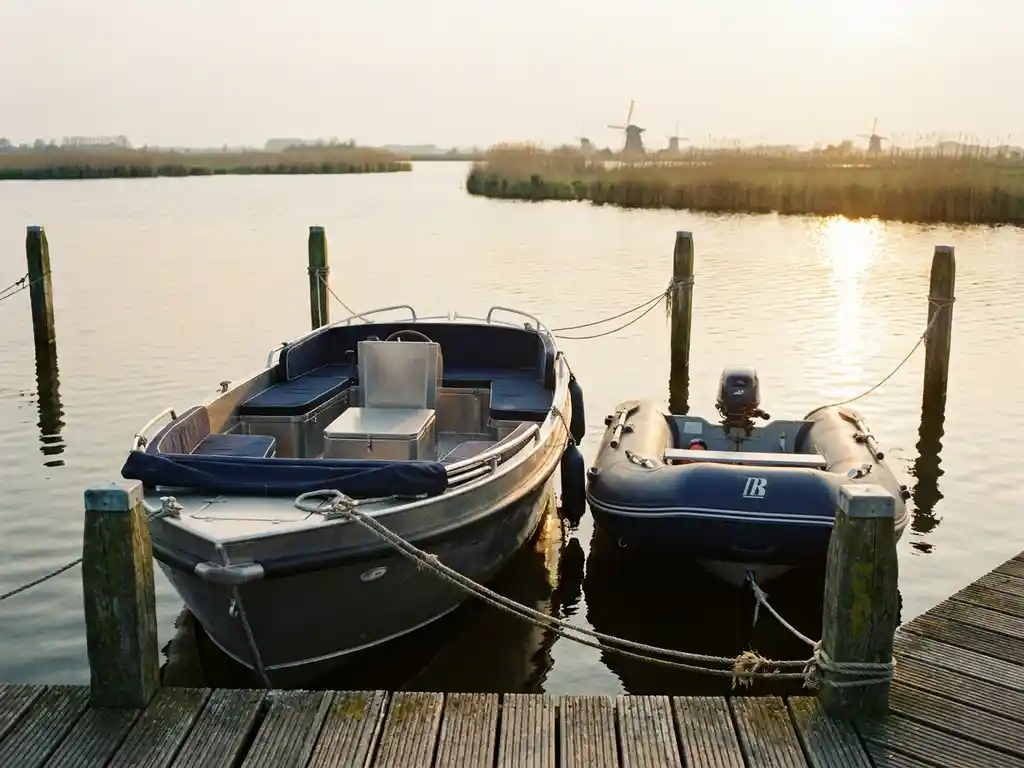 Aluminium sloep en RIB rubberboot afgemeerd aan houten steiger op rustig polderwater, gouden middaglicht weerspiegeld in het water.